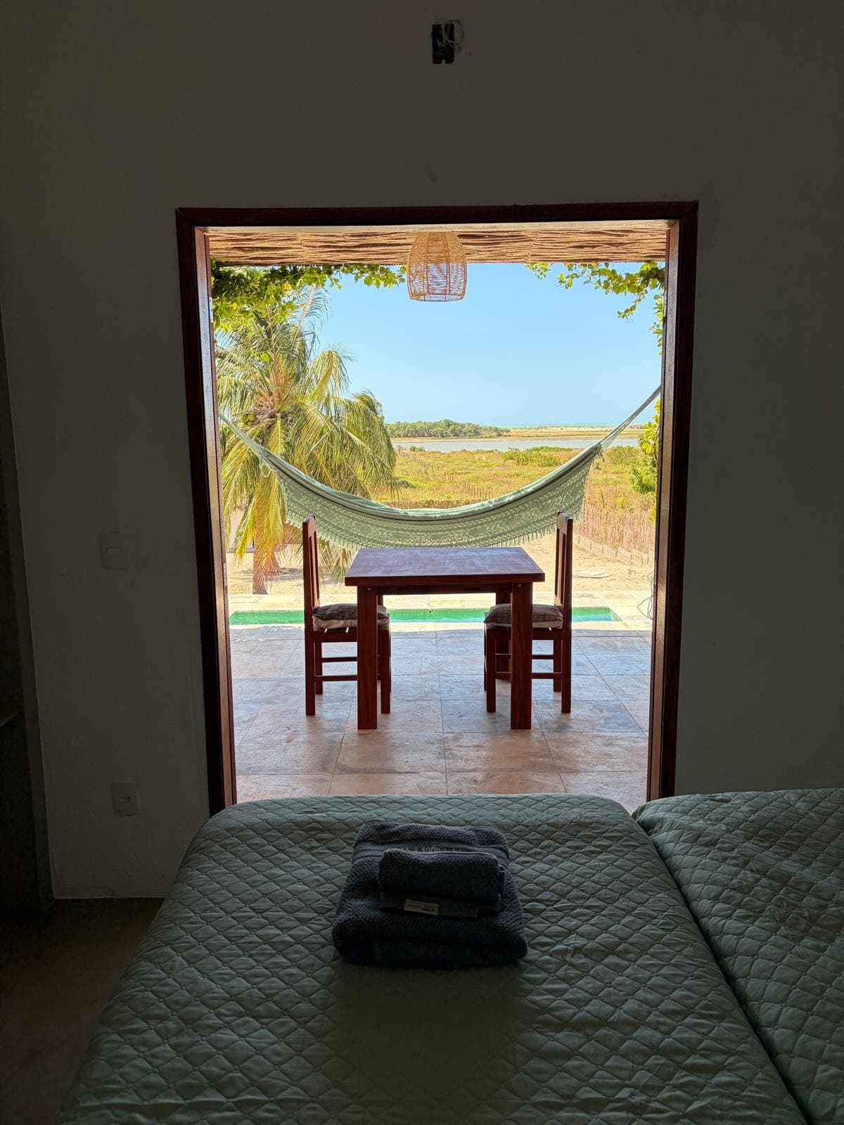 Vue depuis le lit : la porte ouverte sur la terrasse, le hamac et la lagune